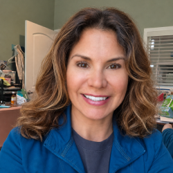 Dr-Sharon-Gold-Team Smiling woman with shoulder-length wavy brown hair in a home or office setting, wearing a blue jacket, photographed indoors with a doorway and window blinds in the background.